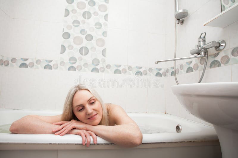 A beautiful smiling girl with blond hair sits in a bath and washes, in the bathroom. An image about cleanliness and about taking care of yourself and your body