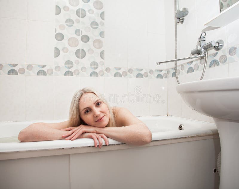 A beautiful smiling girl with blond hair sits in a bath and washes, in the bathroom. An image about cleanliness and about taking care of yourself and your body