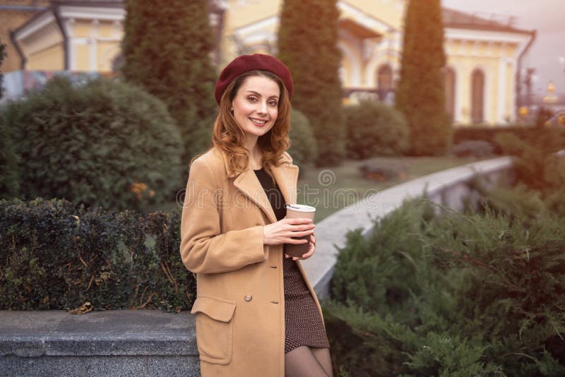 Beautiful smiling Parisian woman holding a paper cup of fresh coffee standing outdoors beautiful garden posing for