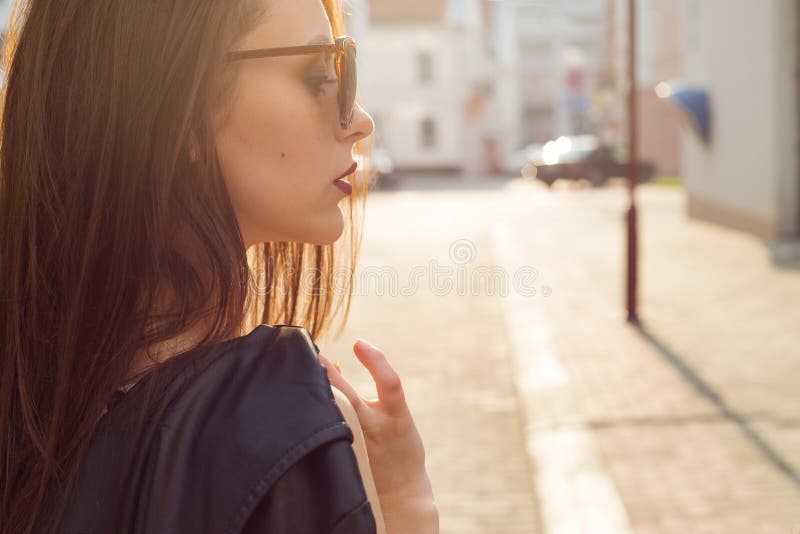 Beautiful stylish girl in a black leather jacket with sunglasses with a dark lipstick in the city at sunset