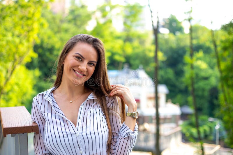 Beautiful successful young woman in white striped shirt, standing outdoors in front of beautiful expensive house surrounded by