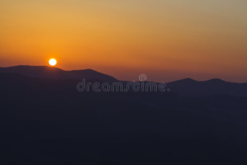 Beautiful sunset in mountains. Wide panorama view of big bright white sun in dramatic orange sky over dark mountain range