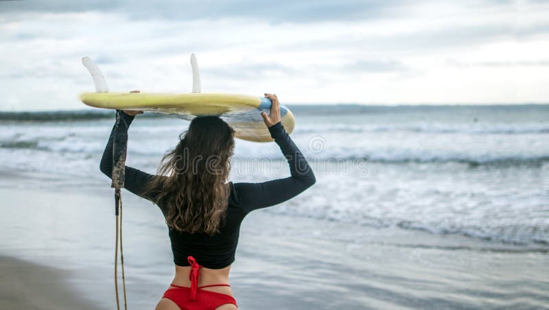 Beautiful surfer girl walking down to the beach for sunset surf session carrying surfboard on head, copy space, fre spce for text