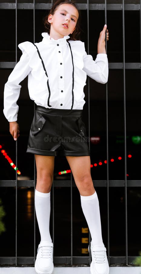 A beautiful and tall schoolgirl in a white blouse, black shorts, socks and sports shoes poses against the background of an iron fence. Vertical photo