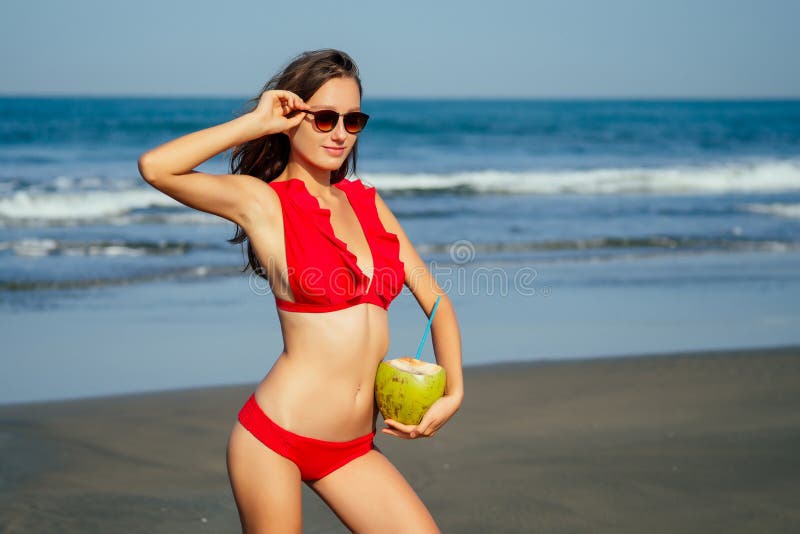 Beautiful tanned girl in trendy red bathing suit sunglasses stands with coconut in her hands on beach tropical island