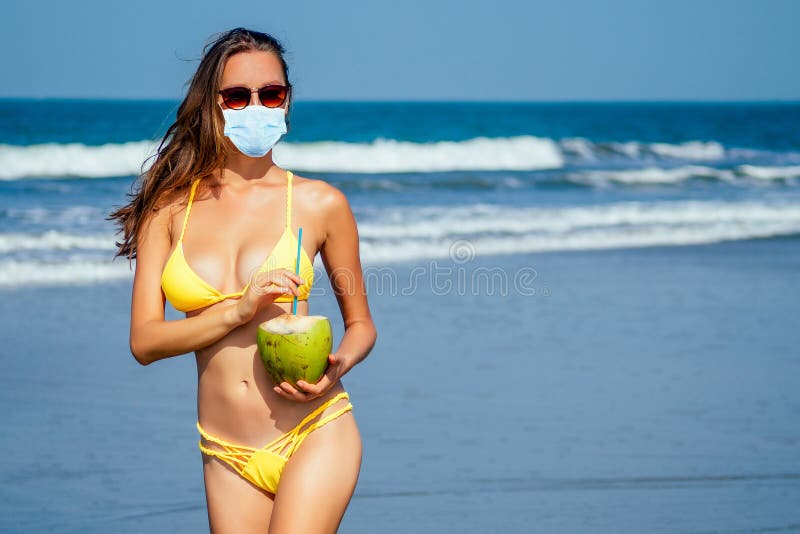 Beautiful tanned girl in trendy yellow bathing suit sunglasses stands with coconut in her hands on beach tropical island