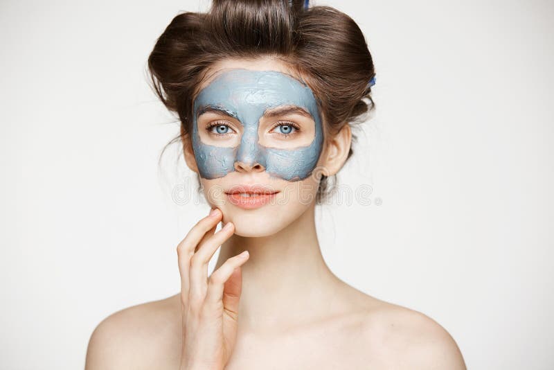 Beautiful tender naked girl in hair curlers and facial mask looking at camera smiling over white background. Beauty