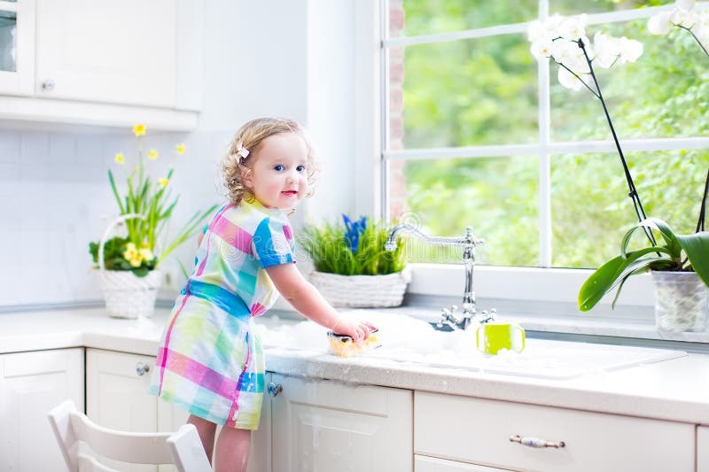 Beautiful toddler girl in colorful dress washing dishes
