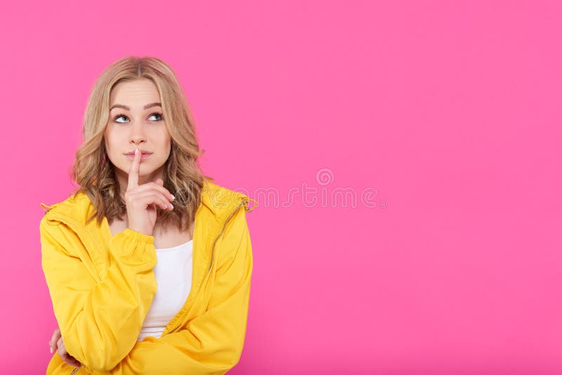 Beautiful trendy girl in colorful clothes deep in thoughts, looking up with finger on lips. Attractive young woman portrait over pastel pink background. Imagination concept. Beautiful trendy girl in colorful clothes deep in thoughts, looking up with finger on lips. Attractive young woman portrait over pastel pink background. Imagination concept.