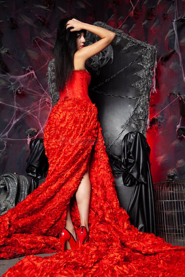 Beautiful vampire woman in red long dress near big black throne in the studio stock photo