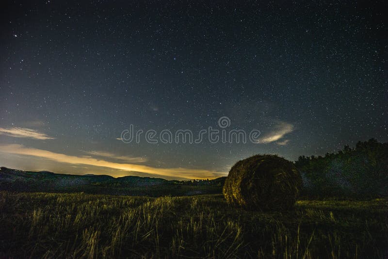 Beautiful view of starred night sky with clouds over a cultivated field with hay bale