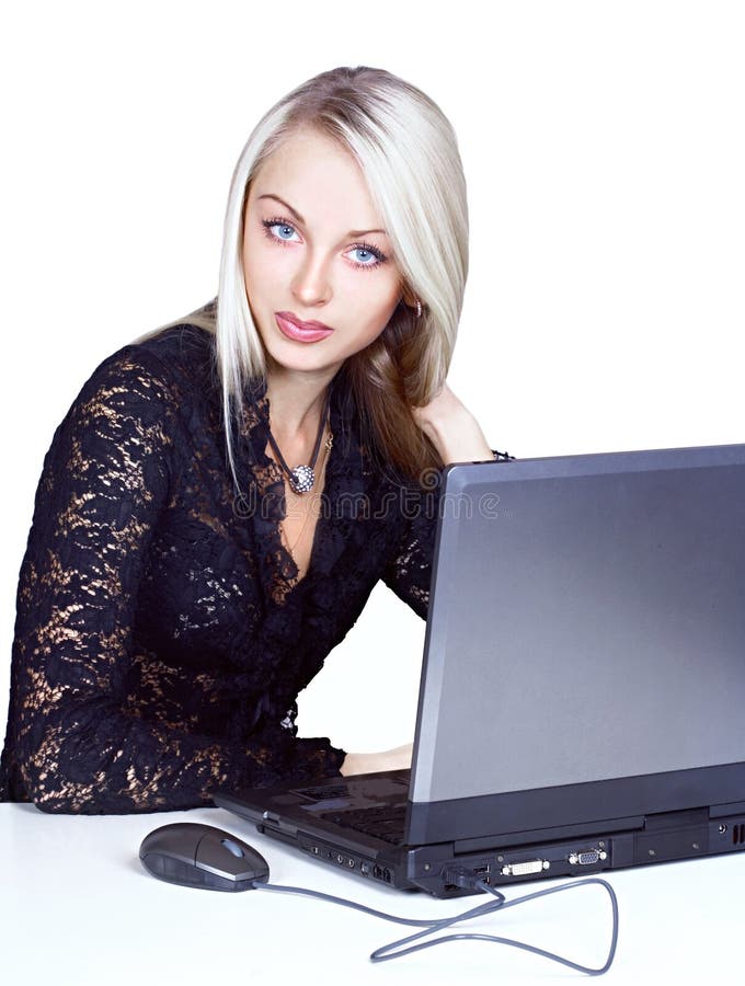 Beautiful blond-haired, blue-eyed woman with a serious expression, wearing a dark lace blouse. An open laptop and mouse are sitting in front of her. Isolated on white background. Beautiful blond-haired, blue-eyed woman with a serious expression, wearing a dark lace blouse. An open laptop and mouse are sitting in front of her. Isolated on white background.