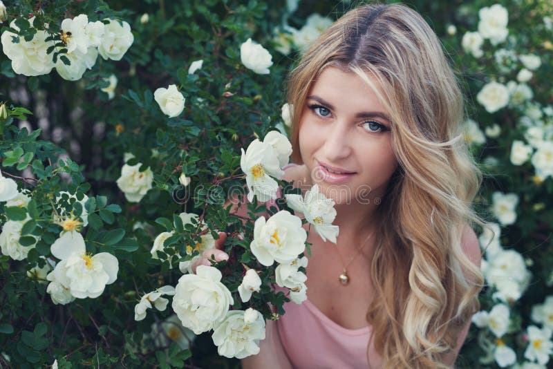 Beautiful woman with long curly hair smells white roses outdoors, closeup portrait of sensual girl face