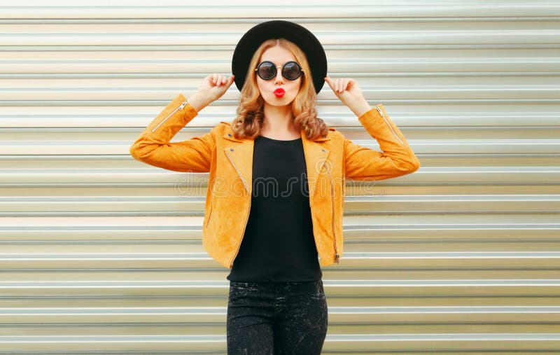 Beautiful woman model blowing red lips sending sweet air kiss wearing yellow jacket, black round hat on wall