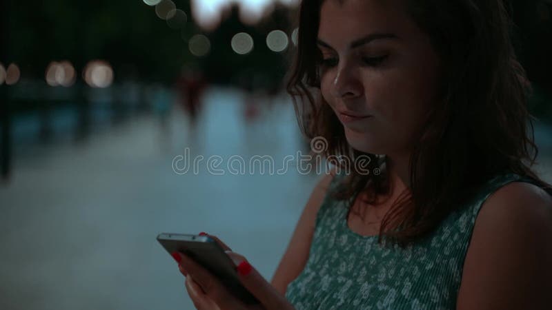 Beautiful Woman with Pink Nails in Blue Dress in the park at dusk chats or texts on smartphone, web scrolling Slow motion