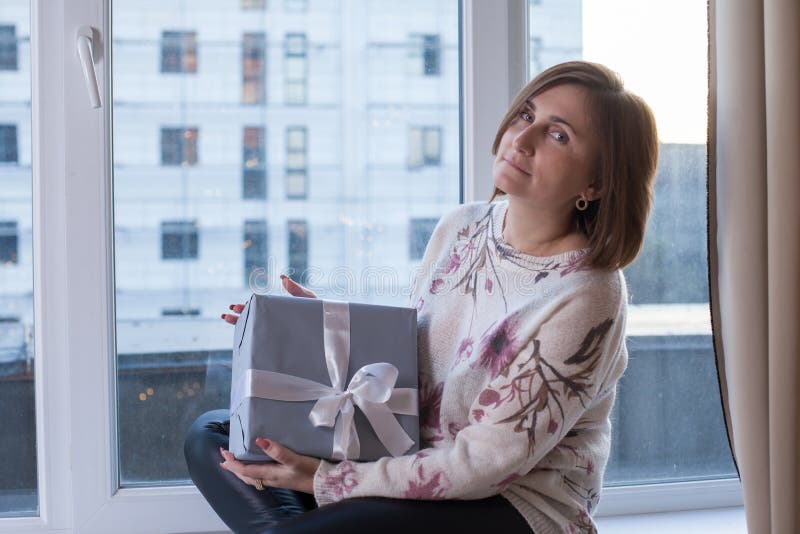 Beautiful woman sitting on windowsill with gift at home stock photo