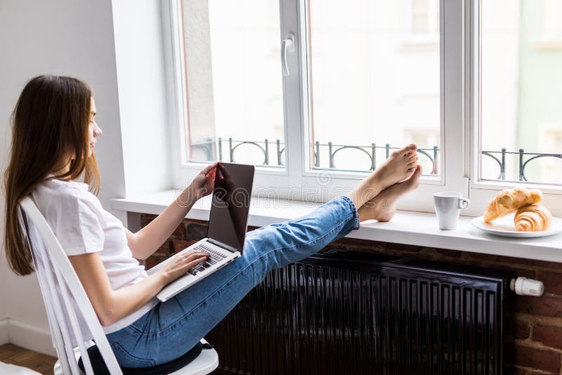 Beautiful woman using a laptop with legs on windowsill and eating breakfast sitting on chair at home stock photo Beautiful woman using a laptop with legs on windowsill and eating breakfast sitting on chair at home stock photo