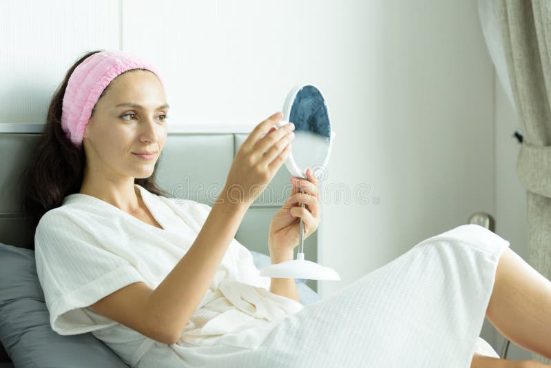 A beautiful woman wearing a towel and a white bathrobe and pink headband has to look a mirror with happy and relaxing on the bed