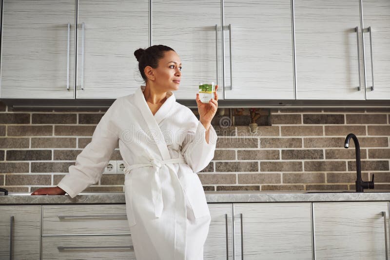 Attractive woman in a white bathrobe is leaning on the kitchen countertop and looking away, drinking lemon water. Concepts of