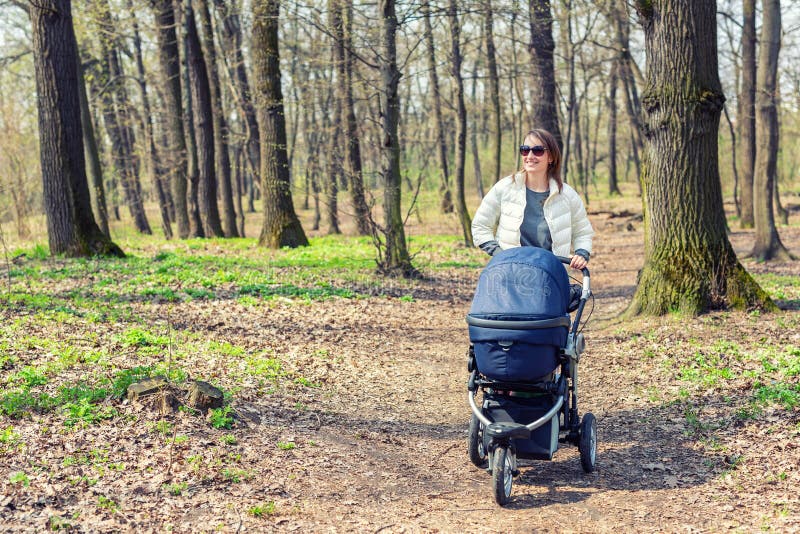 Beautiful young adult woman walking with baby in stroller through forest or park on bright sunny day. Healthy lifestyle royalty free stock image Beautiful young adult woman walking with baby in stroller through forest or park on bright sunny day. Healthy lifestyle royalty free stock image