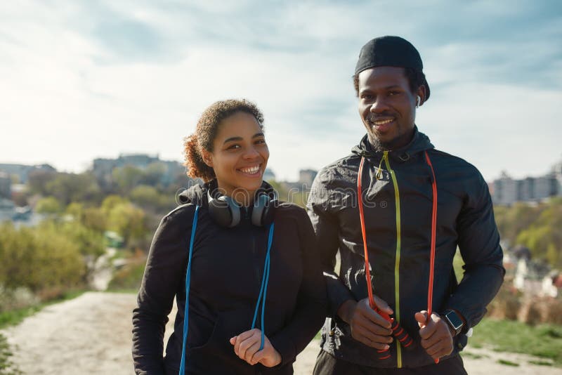 Beautiful young african couple in sportswear holding jumping ropes, looking at camera and smiling while working out together outdoors. Sport, training concept. Healthy active lifestyle