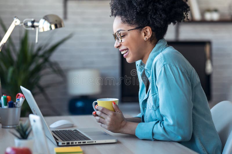 Beautiful young afro business woman making video call with laptop while drinking coffee sitting in the office at home