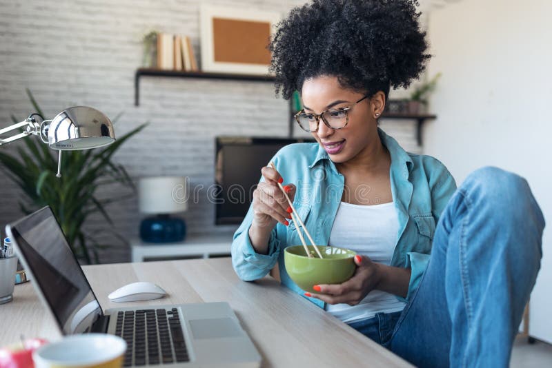 Beautiful young afro business woman making video call with laptop while eating noodles with chopsticks sitting in the office at