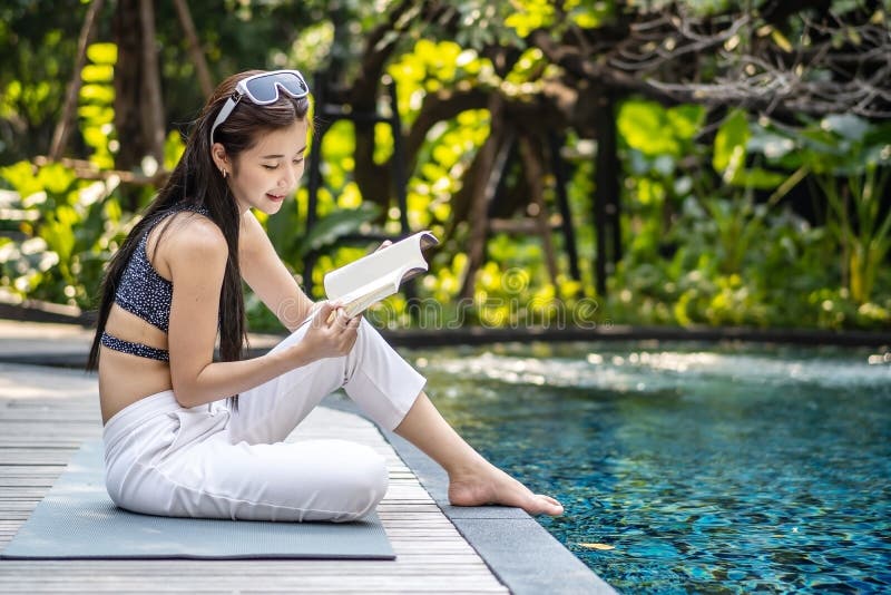 Beautiful young asian woman sits on the pool`s edge outdoors. She reading book while relaxing near swimming pool