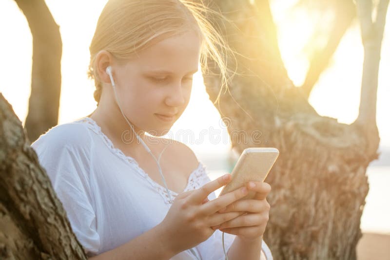 Beautiful young blonde woman listening to music with headphones from a smart phone on nature background, closeup portrait