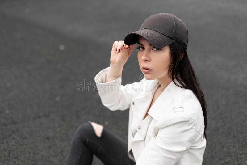 Beautiful young brunette woman with brown eyes in a stylish American baseball cap in a stylish leather white jacket in torn jeans