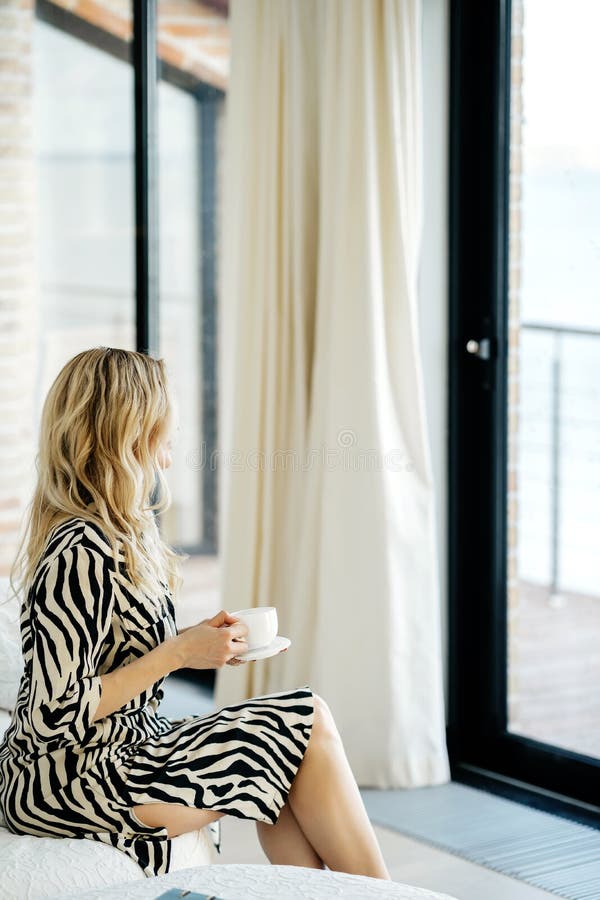 A beautiful young confident and successful girl. With a beautiful make-up and blond wavy-haired sitting on the couch with a cup of coffee. Indoor background royalty free stock images A beautiful young confident and successful girl. With a beautiful make-up and blond wavy-haired sitting on the couch with a cup of coffee. Indoor background royalty free stock images