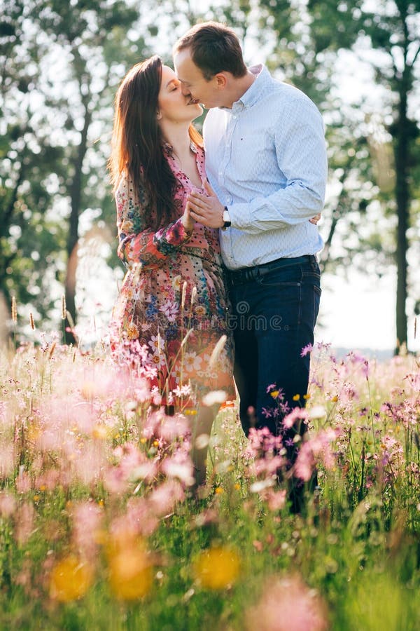 Beautiful young couple holding hands and kissing in sunshine in spring meadow with pink flowers. Happy family embracing in green