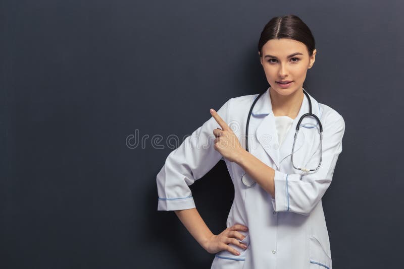 Beautiful young doctor in white medical gown is pointing away and looking at camera, standing against blackboard