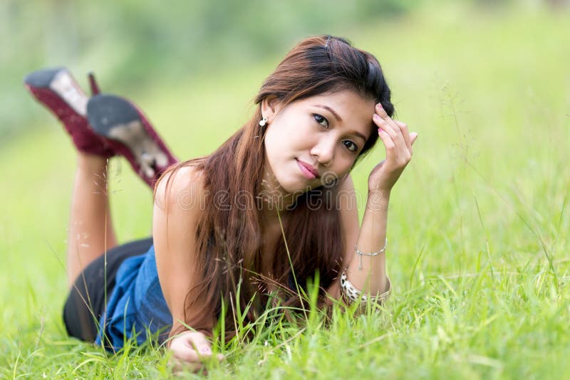 Beautiful young Filipina woman enjoying nature lying on her stomach facing the camera in a green grassy field with a thoughtful expression