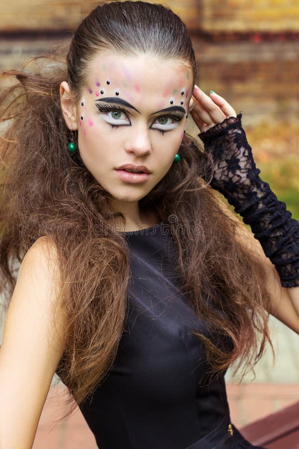 Beautiful young girl on the background of the leaves in autumn day on the street with fantasy makeup in a black dress