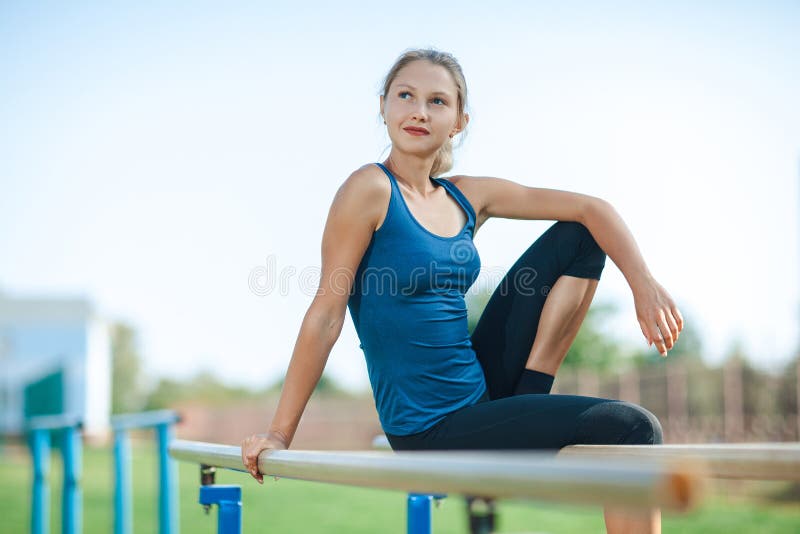 Beautiful young girl in a blue shirt and leggings sitting on the uneven bars, on the outdoor sports ground in summer looking at ca. Mera and smiling stock image Beautiful young girl in a blue shirt and leggings sitting on the uneven bars, on the outdoor sports ground in summer looking at ca. Mera and smiling stock image