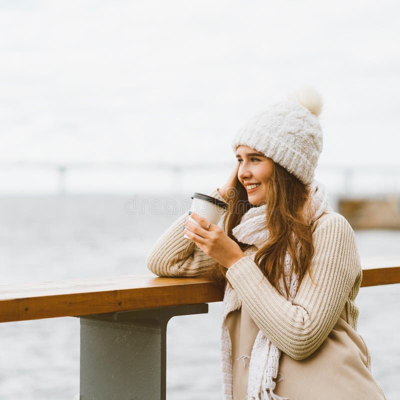 Beautiful young girl drinking coffee, tea from plastic mug in autumn, winter. A woman with long hair stands on waterfront on