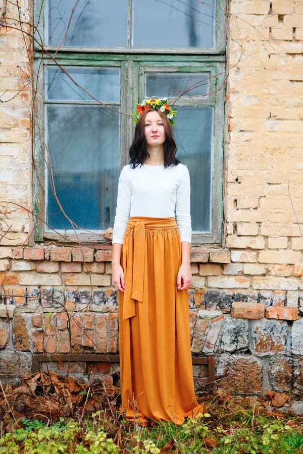 Beautiful young girl with flower wreath on her head near wooden window of an old brick building