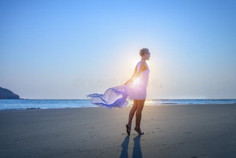 Beautiful young girl with short hair in a long white dress on sandy beach by the sea during sunset. Counter light stock images Beautiful young girl with short hair in a long white dress on sandy beach by the sea during sunset. Counter light stock images