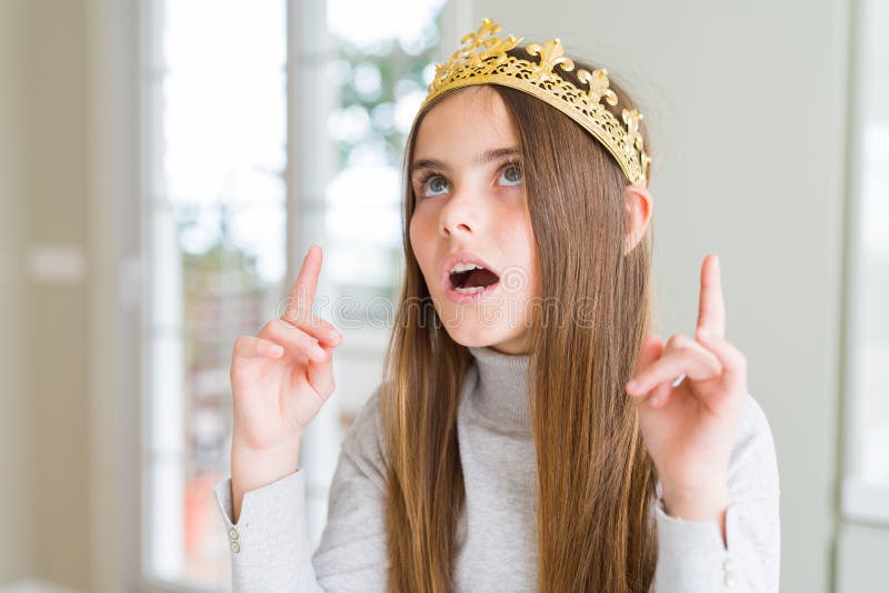 Beautiful young girl wearing a golden crown as a princess from fairytale amazed and surprised looking up and pointing with fingers