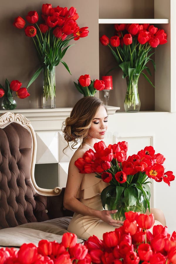 A beautiful young girl in a white dress is sitting on the sofa and holding a large bouquet of red tulips. March 8 concept. Morning of the bride