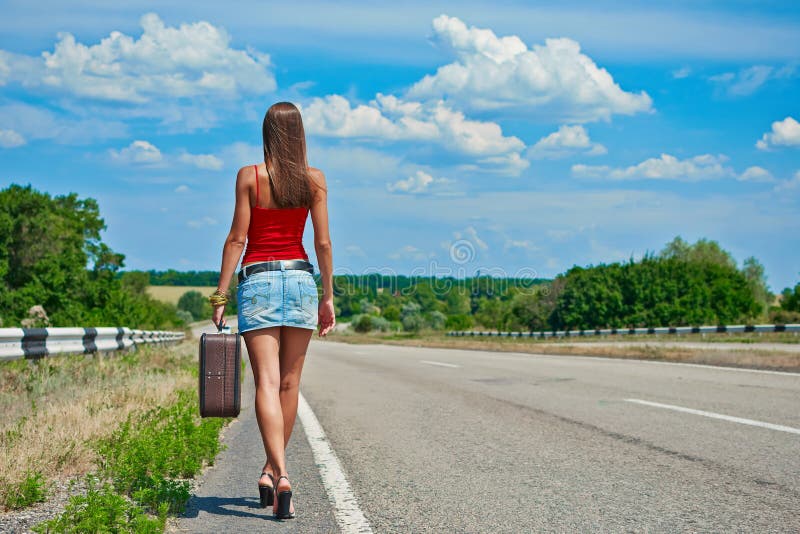 Beautiful young girl or woman in mini with suitcase hitchhiking along a road