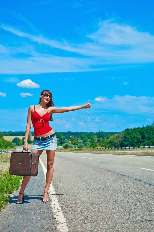 Beautiful young girl or woman in mini with suitcase hitchhiking along a road