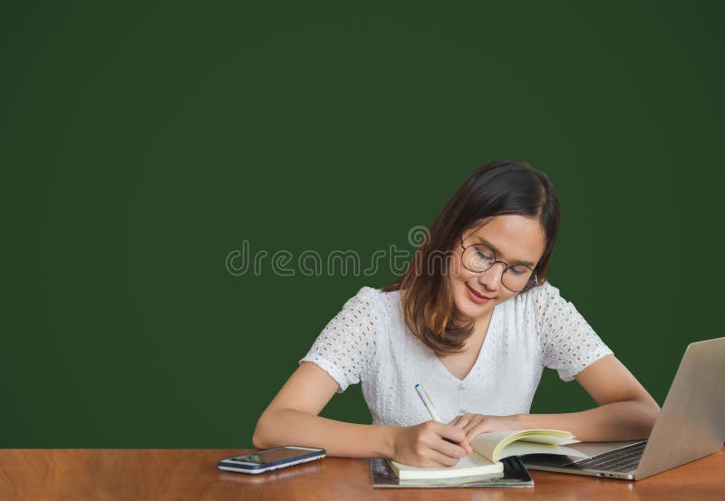 Beautiful young glasses Asian woman sitting and writing on  a book with laptop at side with green background