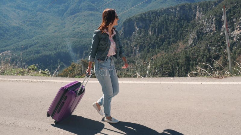 Beautiful young lady on rural road with suitcase hitchhiking on sunny day outdoors