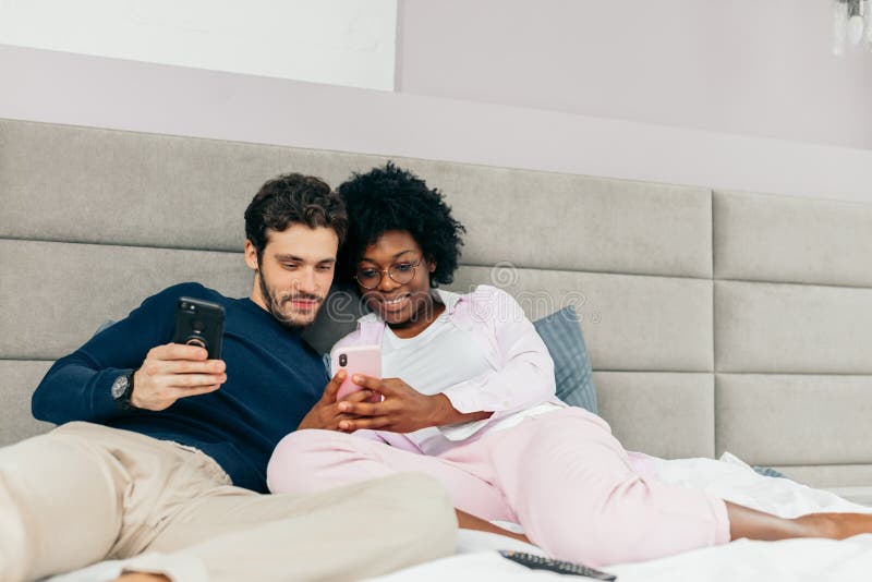 Young wife and husband relaxing on bed during their day-off. Spauses of different nationalities making selfie or watching videos using smart phones. Young wife and husband relaxing on bed during their day-off. Spauses of different nationalities making selfie or watching videos using smart phones