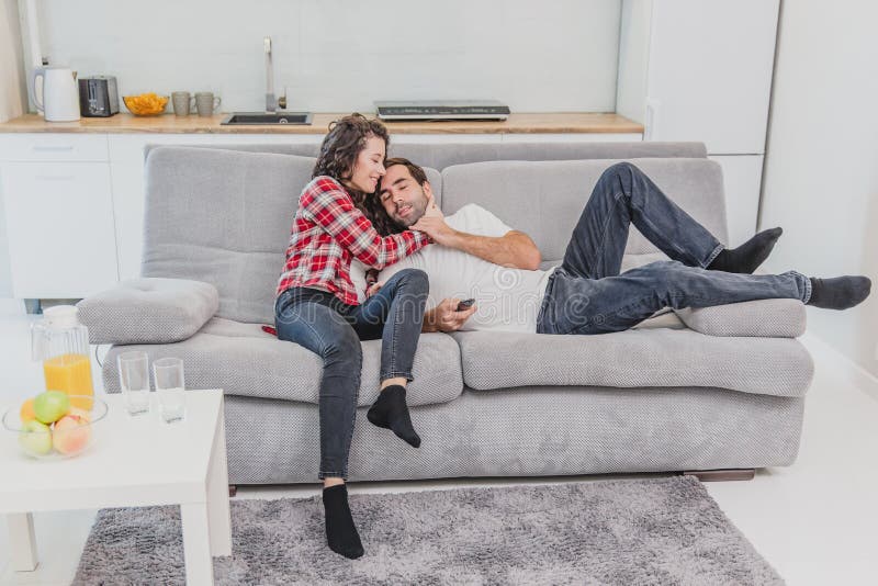 Beautiful young married couple is watching tv with joy. They are sitting on sofa and embracing. The men is holding the remote. They are smiling. Beautiful young married couple is watching tv with joy. They are sitting on sofa and embracing. The men is holding the remote. They are smiling