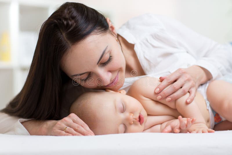 Beautiful young mom looking at her sleeping little baby and smiling. Child lying in bed at home royalty free stock photography Beautiful young mom looking at her sleeping little baby and smiling. Child lying in bed at home royalty free stock photography