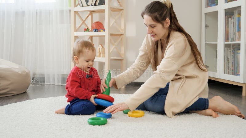 Beautiful young mother playing with little baby son on floor and assembling toy tower. Baby development, child playing games
