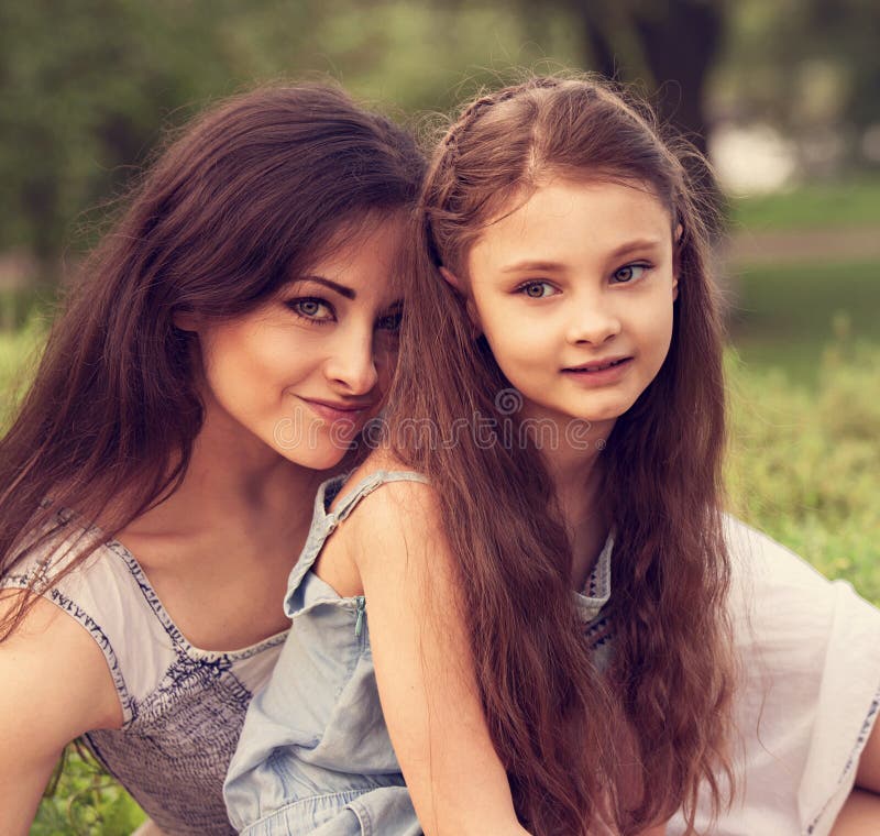 Beautiful young natural mother embracing her cute long hair daughter on summer green grass background in sunny day. Portrait of